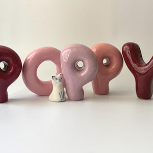 Könnte beinhalten: Keramikskulptur mit dem Wort "POPPY". Die Buchstaben sind in Rosa- und Burgundertönen gehalten, mit einer kleinen weißen und blauen Katzenfigur zwischen dem "O" und dem "P".