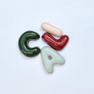 May include: Four glazed ceramic shapes: a dark green "C", a light pink oblong, a deep red heart, and a light grey "A" with a hole. The shapes are on a white background.