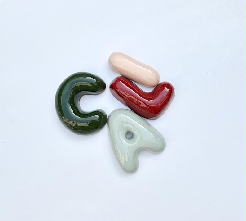May include: Four glazed ceramic shapes: a dark green "C", a light pink oblong, a deep red heart, and a light grey "A" with a hole. The shapes are on a white background.