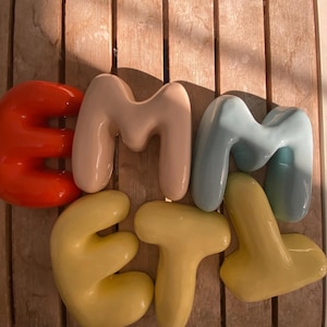 May include: A collection of colorful, glossy ceramic letters spelling out "EMMET". The letters are in shades of red, beige, light blue, and yellow, arranged on a wooden surface.