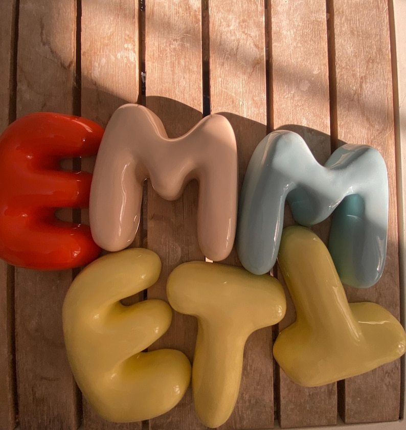 May include: A set of glossy ceramic letters spelling "EMMET". The letters are in red, beige, light blue, and yellow, arranged on a wooden surface.
