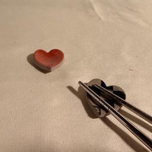 Op de afbeelding: Twee hartvormige keramische houders. Eén is effen matrood van kleur. De andere is zwart-wit met een geblokt patroon en ondersteunt een paar metalen zilverkleurige eetstokjes die op het holle oppervlak rusten.