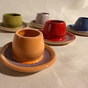 May include: A set of five ceramic egg cups, each featuring a smooth, solid-colored cup in orange, red, green, blue, or light purple, resting on a matching saucer with a white and dark purple honeycomb pattern on the interior surface.