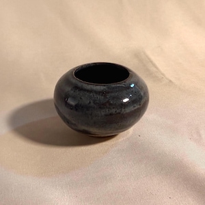 May include: A small, round ceramic bowl with a glossy finish. The exterior features a dark, mottled glaze with blended shades of deep blue, charcoal gray, and hints of brown. The interior of the bowl is dark, and the rim is smooth and circular.