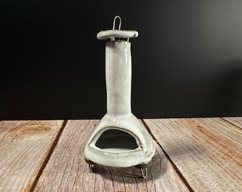 Puede incluir: Una chimenea de cerámica en un esmalte gris claro con una pequeña abertura y un asa en la parte superior. La chimenea tiene una base redondeada y se apoya en tres pequeños pies metálicos. El fondo es oscuro y la chimenea está sobre una superficie de madera.