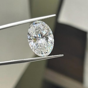 May include: An oval-cut diamond held by tweezers. The diamond is clear and reflects light, showcasing its facets. The background is blurred, emphasising the diamond's brilliance and clarity. The tweezers are silver.