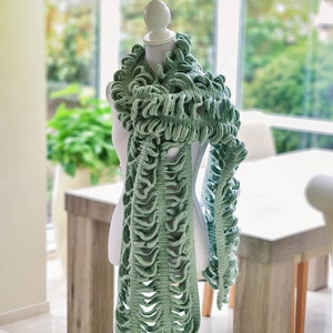 May include: A green crocheted scarf with a ruffled design is draped around a white mannequin. The scarf is made with a delicate openwork stitch.