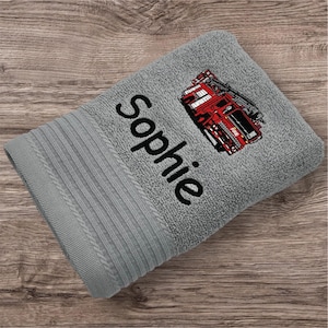 Könnte beinhalten: Ein graues Badetuch mit dem Namen "Sophie" in schwarzer Schrift gestickt. Ein rotes Feuerwehrauto ist auf das Handtuch gestickt. Das Handtuch ist gefaltet und hat eine gerippte Bordüre.