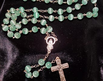 May include: A rosary featuring light green, spherical beads and silver-coloured chain links. It includes a silver crucifix and a religious figure centerpiece. The beads have a cat's eye effect.