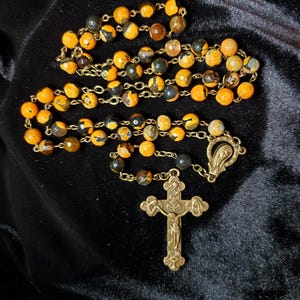 Könnte beinhalten: Ein Rosenkranz mit orangefarbenen und schwarzen gesprenkelten Perlen und einem goldfarbenen Metallkreuz. Die Perlen sind auf einer goldfarbenen Kette aufgereiht. Das Kreuz hat ein verziertes Design.