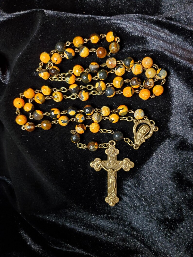 K&ouml;nnte beinhalten: Ein Rosenkranz mit orangefarbenen und schwarzen gesprenkelten Perlen und einem goldfarbenen Metallkreuz. Die Perlen sind auf einer goldfarbenen Kette aufgereiht. Das Kreuz hat ein verziertes Design.