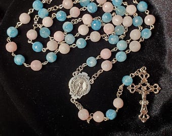 May include: A rosary featuring pink, blue, and white gemstone beads. The beads are strung on a silver chain, with a silver crucifix and a central medallion. The rosary's design is delicate, with a mix of colours and textures.