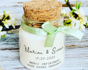 May include: A small glass jar candle with a cork stopper and a light green ribbon tied around the neck. The label features the text Marine & Simon, the date 17.07.2020, and the French phrase MERCI INFINIMENT POUR VOTRE PRÉSENCE in black ink.