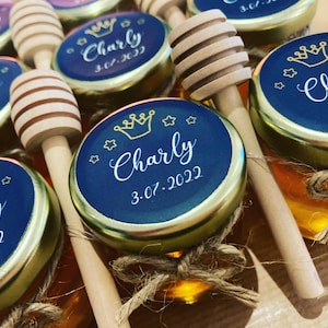 Peut inclure: Petits pots en verre remplis de miel doré, chacun doté d'une étiquette circulaire bleu foncé avec une icône de couronne dorée, des étoiles, le nom Charly et la date 3.07.2022. Une cuillère à miel en bois est attachée à chaque pot avec de la ficelle.