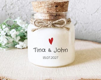 May include: A clear glass jar candle with a cork stopper and a jute twine bow tied around the neck. The front of the candle features a small red heart, the names Tina & John, and the date 15.07.2027 printed in black text on the white wax.