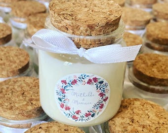 May include: A small glass jar filled with cream-colored wax, topped with a natural cork stopper and tied with a white ribbon. A circular label features a floral wreath design in pink and blue with the text Mathilde & Maxence printed in the center.