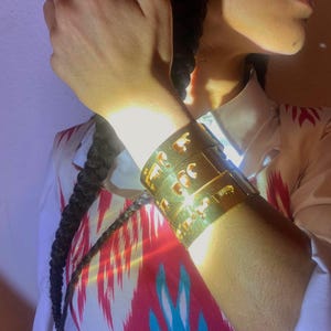 May include: A wide gold-toned metal cuff bracelet featuring intricate cut-out designs. The bracelet has a polished finish and is worn on the wrist, displaying a series of repeating geometric and organic shapes carved into the surface of the metal.