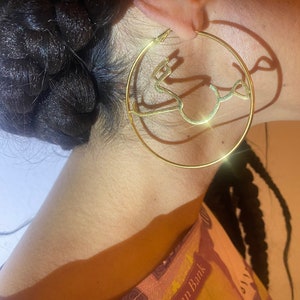 May include: Gold hoop earrings with a decorative design in the shape of a stylized letter. The earrings are large and have a shiny finish.