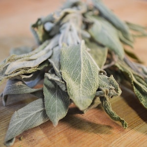 May include: A bundle of fresh sage leaves, with a soft, textured appearance, rests on a wooden surface. The sage leaves are a muted green color, with a slightly fuzzy texture. The image is a close-up, highlighting the details of the leaves.