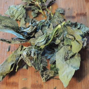 May include: Dried green leaves with stems, possibly herbs or spices, are arranged on a wooden cutting board. The leaves have a wrinkled texture and varying shades of green and brown. The image is well-lit, highlighting the natural colors and textures.