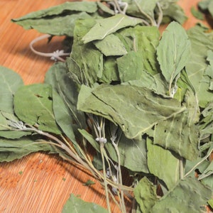 Puede incluir: Hojas verdes secas, posiblemente hierbas o especias, dispuestas sobre una superficie de madera marr&oacute;n claro. Las hojas tienen un aspecto ligeramente descolorido, lo que sugiere que est&aacute;n secas.