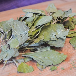 Puede incluir: Primer plano de hojas verdes secas, posiblemente hierbas o especias, dispuestas sobre una tabla de cortar de madera. Las hojas tienen un aspecto descolorido, con venas visibles. Fondo de tela morada.