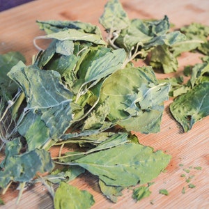 Puede incluir: Primer plano de verduras de hojas verdes frescas sobre una tabla de cortar de madera. Las hojas tienen un aspecto ligeramente seco, con venas y bordes visibles. La tabla de cortar es de madera marr&oacute;n claro.