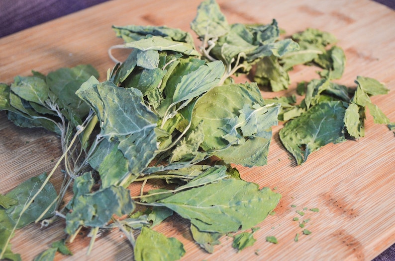 Puede incluir: Primer plano de verduras de hojas verdes frescas sobre una tabla de cortar de madera. Las hojas tienen un aspecto ligeramente seco, con venas y bordes visibles. La tabla de cortar es de madera marr&oacute;n claro.