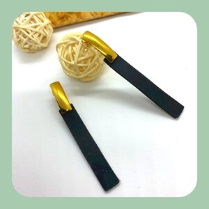 Puede incluir: Un par de pendientes de cuero negro con detalles dorados. Los pendientes son de forma rectangular y tienen un diseño simple y minimalista.