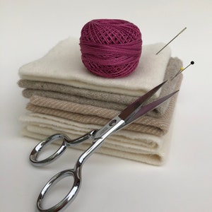 Puede incluir: Una pila de cuadrados de tela blanca y beige, una madeja de hilo rosa, un par de tijeras plateadas y dos alfileres.