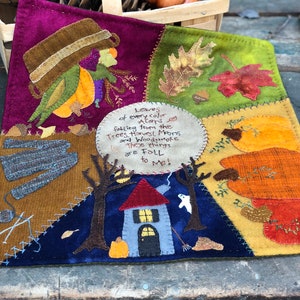 May include: A colorful felt wall hanging with a stitched design of a house, trees, pumpkins, leaves, and acorns. The text reads "Leaves of every color Acorns falling from the Trees, Harvest Moons and Woodsmoke These things are FOLL to me!"