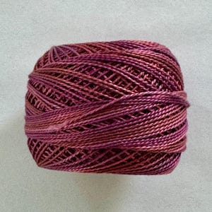 May include: A ball of variegated embroidery floss in shades of burgundy, rose, and peach. The thread is tightly wound, showing the texture of the fibers. Ideal for embroidery, cross-stitch, or other needlework projects.