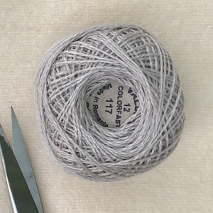 Op de afbeelding: Een bol lichtgrijze borduurgaren met een wit label met de tekst "COLORFAST 12 117 Gemaakt in Roemenië"