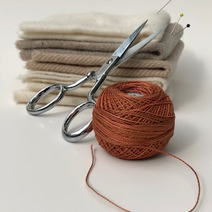 May include: A ball of orange embroidery floss with a length of thread extending from it, a pair of silver scissors, and a stack of beige and white fabric swatches.