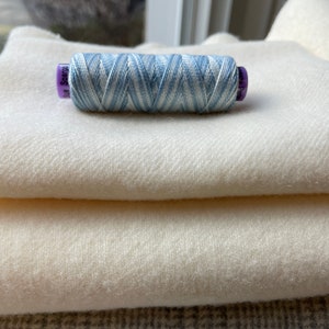 May include: A spool of blue and white striped thread with the text "Sue Spargo" on the label. The thread is sitting on top of a stack of three white fabric swatches.