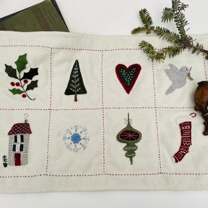 Op de afbeelding: Een witte vilt kerst-adventskalender met negen geborduurde vierkanten. Elk vierkant heeft een ander kerstthema, waaronder een hulsttak, een kerstboom, een hart, een duif, een huis, een sneeuwvlok, een ornament en een sok.