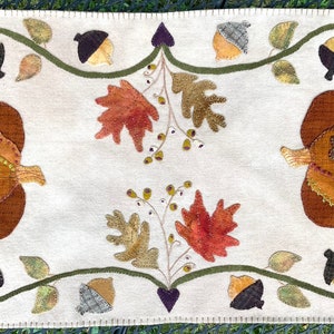 May include: A cream-colored rectangular table runner with autumn-themed applique. The design features two pumpkins, leaves, and acorns in shades of brown, orange, and green. The edges are finished with a stitched border.