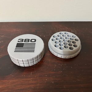 May include: A light gray 380 ammunition container with a lid featuring the number "380" and a flag design. The container is cylindrical with a grid-like pattern on the sides. The open section has multiple circular holes.