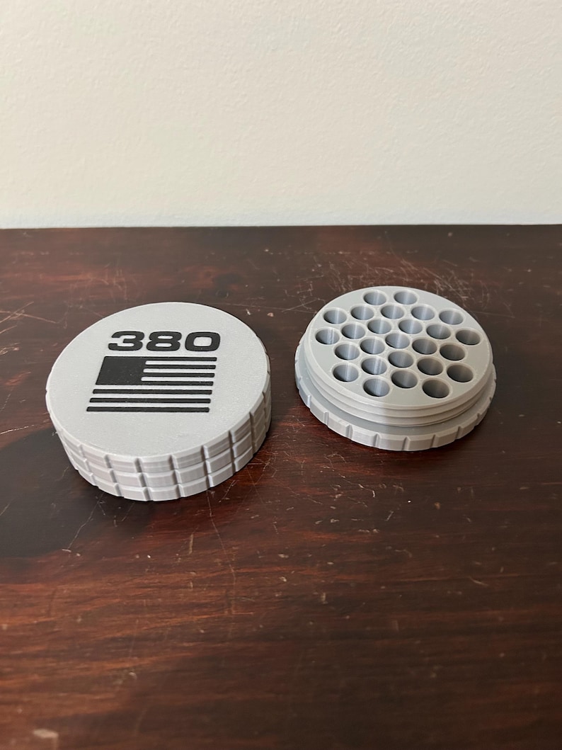 May include: A light gray 380 ammunition container with a lid featuring the number "380" and a flag design. The container is cylindrical with a grid-like pattern on the sides. The open section has multiple circular holes.