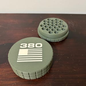 May include: An olive green container with a lid featuring the number "380" and a white American flag design. The container has a ridged edge and a circular top with multiple holes. The second piece is a matching olive green circular piece with multiple holes.
