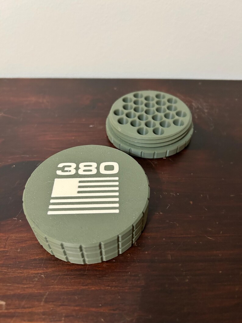 May include: An olive green container with a lid featuring the number "380" and a white American flag design. The container has a ridged edge and a circular top with multiple holes. The second piece is a matching olive green circular piece with multiple holes.