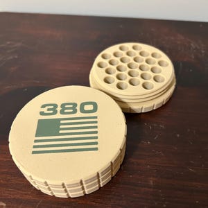 May include: Tan-colored round container with the number "380" and a green flag design. The container is open, revealing a grid of small holes. The container is made of a ridged material.