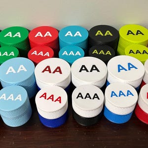 May include: A collection of colorful cylindrical objects with textured sides and flat tops. Each top features the letters "AA" or "AAA" in a contrasting color. Colors include blue, green, red, black, yellow, pink, brown, and white.