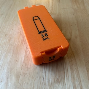 K&ouml;nnte beinhalten: Eine orangefarbene rechteckige H&uuml;lle mit einer schwarzen Umrisszeichnung einer Kugel und dem Text "38 SPL". Die H&uuml;lle ist geschlossen und hat einen Klappdeckel. Die H&uuml;lle liegt auf einer Holzoberfl&auml;che.
