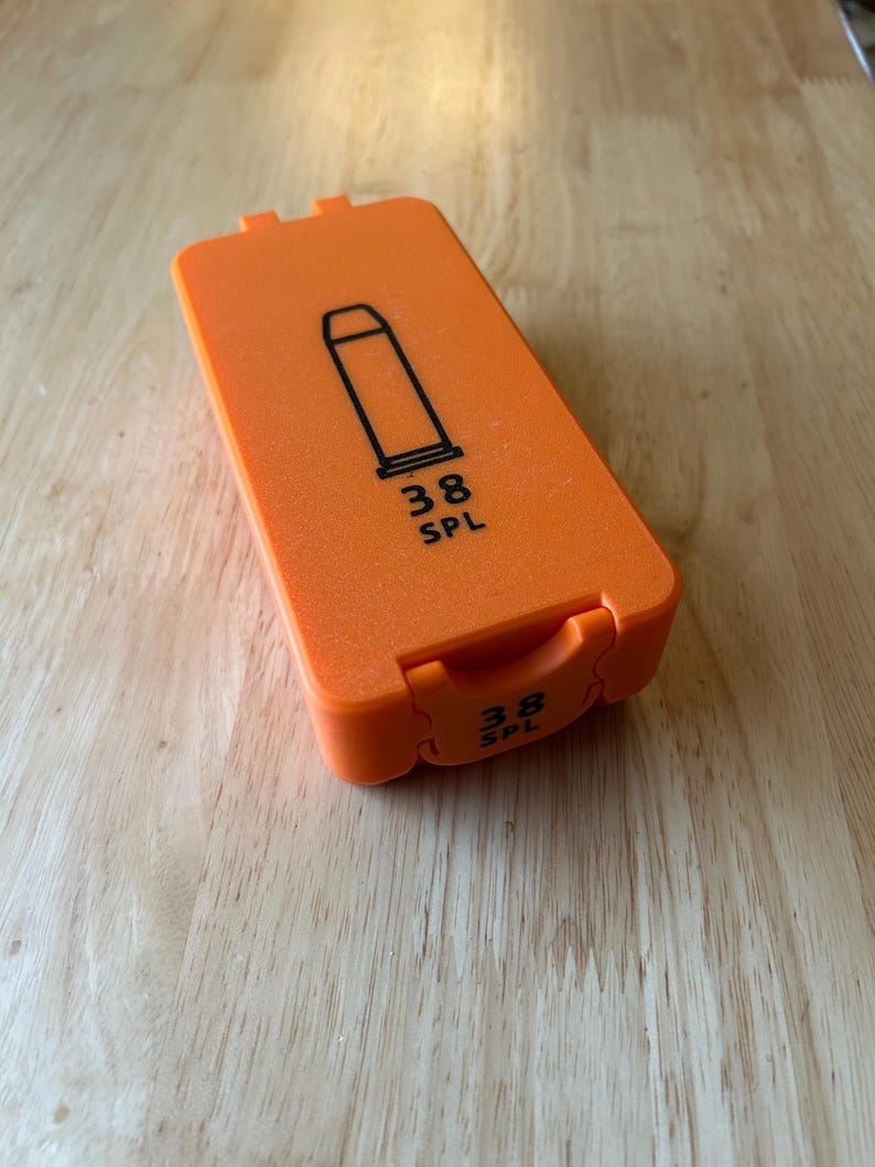 K&ouml;nnte beinhalten: Eine orangefarbene rechteckige H&uuml;lle mit einer schwarzen Umrisszeichnung einer Kugel und dem Text "38 SPL". Die H&uuml;lle ist geschlossen und hat einen Klappdeckel. Die H&uuml;lle liegt auf einer Holzoberfl&auml;che.