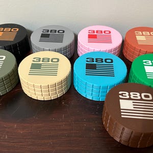 May include: A collection of eight round, colorful objects with a textured, brick-like pattern on the sides. Each object has a different color top, with the number "380" and a stylized American flag design printed on top.