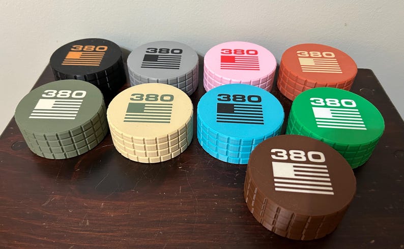 May include: A collection of eight round, colorful objects with a textured, brick-like pattern on the sides. Each object has a different color top, with the number "380" and a stylized American flag design printed on top.
