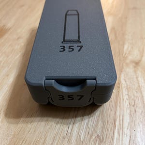 May include: A gray plastic ammunition case with a hinged lid. The case features a black outline of a bullet and the number "357" printed on the top and bottom. The case is rectangular and has a textured surface.