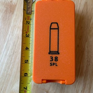 K&ouml;nnte beinhalten: Ein orangefarbenes rechteckiges Etui mit einer schwarzen Umrisszeichnung einer Patrone und dem Text "38 SPL". Das Etui ist etwa 18 cm lang. Ein gelbes Ma&szlig;band liegt daneben.