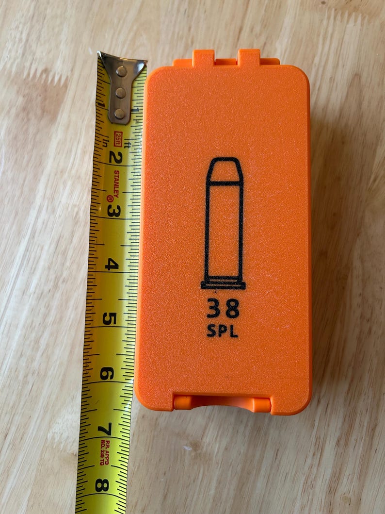 K&ouml;nnte beinhalten: Ein orangefarbenes rechteckiges Etui mit einer schwarzen Umrisszeichnung einer Patrone und dem Text "38 SPL". Das Etui ist etwa 18 cm lang. Ein gelbes Ma&szlig;band liegt daneben.